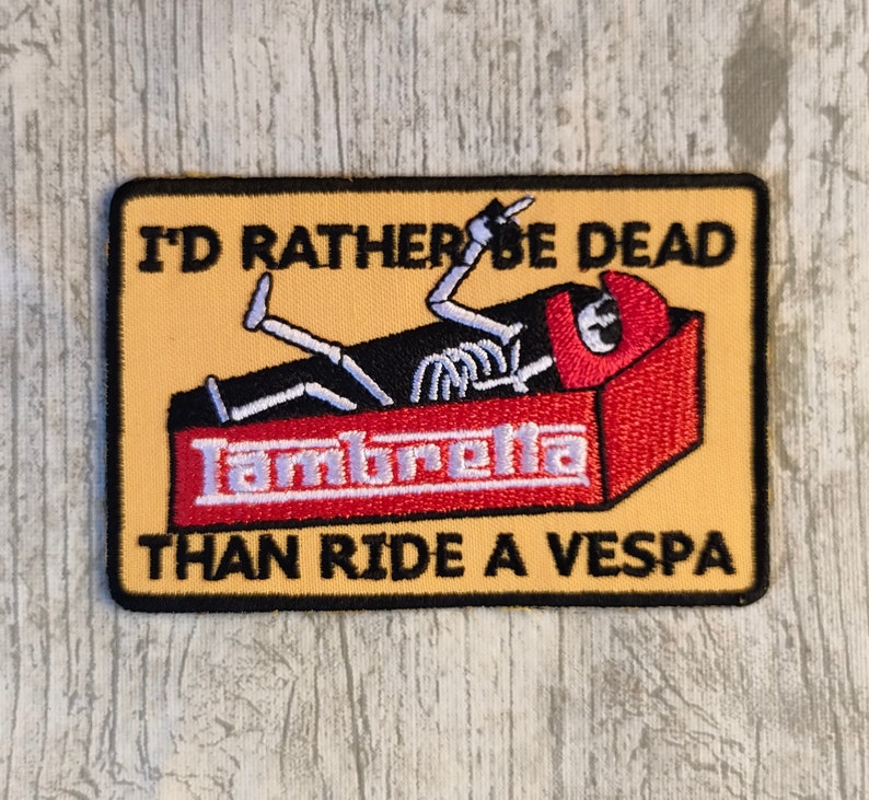 May include: A yellow and black embroidered patch featuring a skeleton in a coffin with the text "I'd rather be dead than ride a Vespa" and "Lambretta" on the coffin.