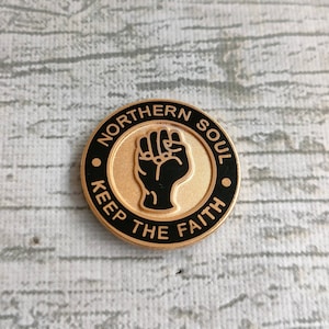 Può includere: Uno spillo dorato con contorno nero. Lo spillo presenta un pugno nero in rilievo e il testo "NORTHERN SOUL KEEP THE FAITH".