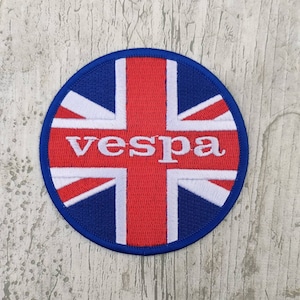 Puede incluir: Parche redondo bordado con el diseño de la bandera de la Union Jack en rojo, blanco y azul. La palabra "vespa" está bordada en blanco en el centro. El parche tiene un borde azul, ideal para ropa o accesorios.