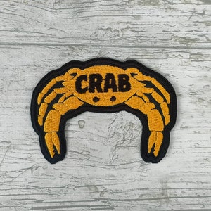 May include: Embroidered patch with a crab design in yellow and black. The word "CRAB" is embroidered in black across the crab's body. The patch has a black border, suitable for application on clothing or accessories.