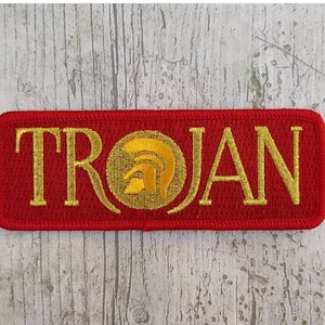 Puede incluir: Parche bordado rojo con la palabra "TROJAN" en letras doradas y un gráfico de casco dorado.