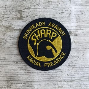 Könnte beinhalten: Runder, gestickter Aufnäher, marineblau mit goldenem Text. Der Text lautet "SKINHEADS AGAINST RACIAL PREJUDICE" und zeigt das Wort "SHARP" in einer markanten Schriftart innerhalb eines goldenen Kreises.
