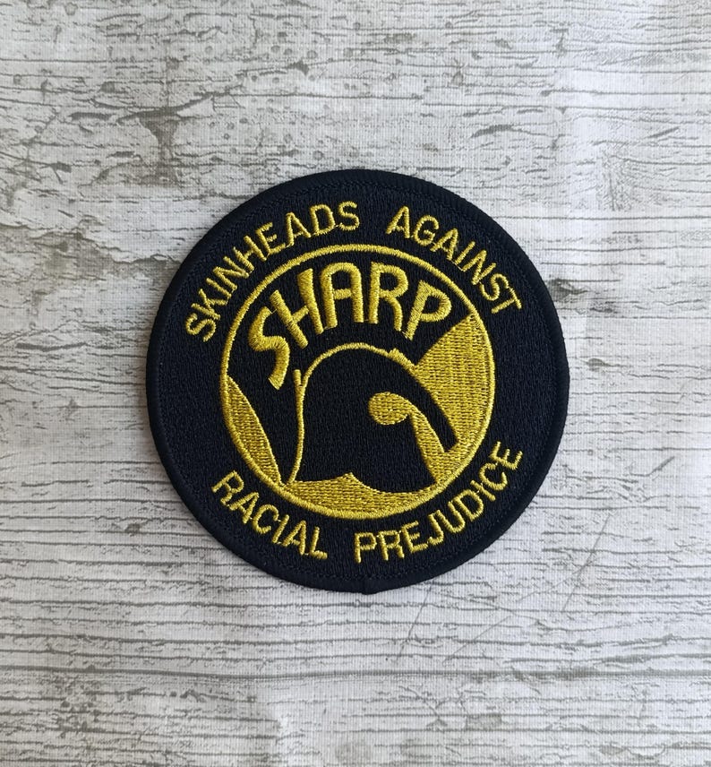 May include: Round, embroidered patch with the text "SKINHEADS AGAINST RACIAL PREJUDICE" in gold thread on a navy blue background. The word "SHARP" is in a stylized font within a gold circle.