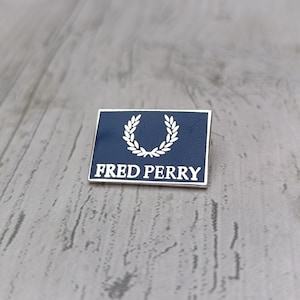 May include: A navy blue rectangular pin with a silver border. It displays a white laurel wreath and the words "FRED PERRY" in white. The pin is set on a wooden surface. The design is simple and classic.