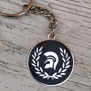 Può includere: Un portachiavi nero e bianco con un logo mod a bersaglio con un casco bianco e una corona di alloro.