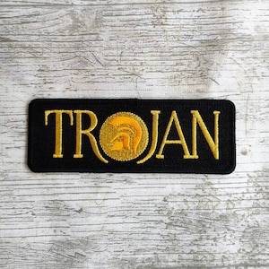 May include: Black embroidered patch with the word "TROJAN" in gold lettering and a gold helmet graphic.
