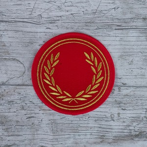 May include: A round, red patch with a gold laurel wreath design. The wreath encircles a blank red center. The patch is likely an emblem or decorative element, possibly for clothing or accessories.