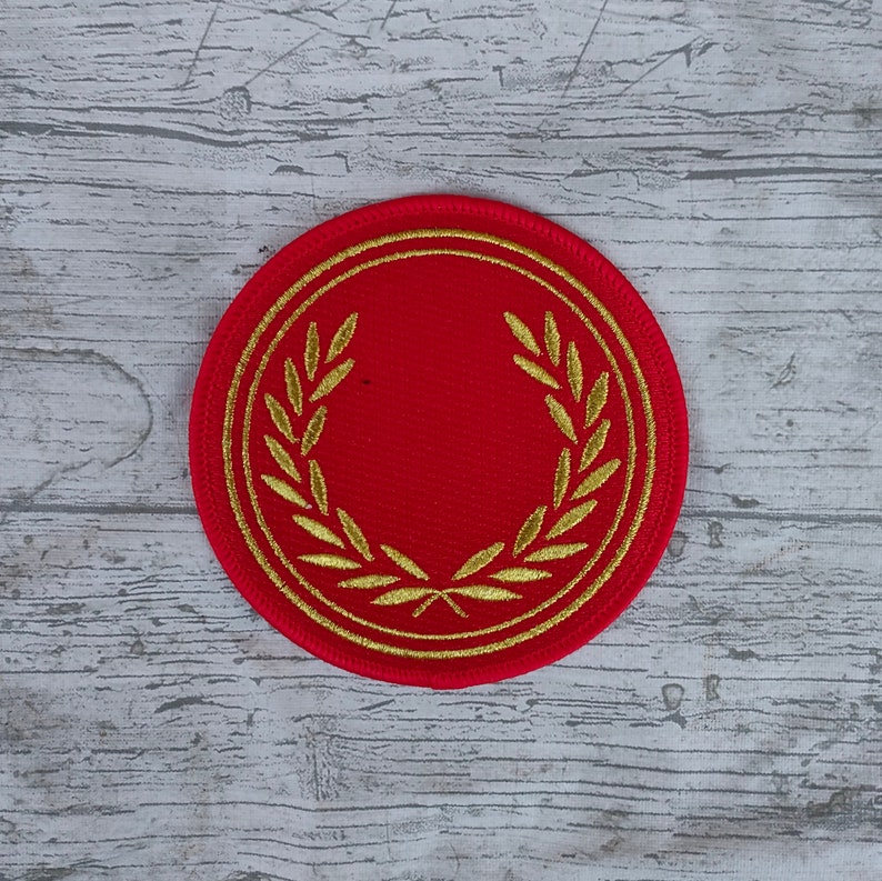 K&ouml;nnte beinhalten: Ein runder, roter Aufn&auml;her mit einem goldenen Lorbeerkranz-Design. Der Kranz umschlie&szlig;t eine leere rote Mitte. Der Aufn&auml;her ist wahrscheinlich ein Emblem oder ein dekoratives Element, m&ouml;glicherweise f&uuml;r Kleidung oder Accessoires.