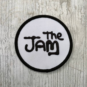 Puede incluir: Un parche circular blanco con un borde negro. El parche tiene texto bordado en negro que dice "The Jam" en una fuente estilizada.