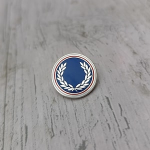 Könnte beinhalten: Runder, silberfarbener Ansteckpin mit blauem Zentrum und weißem Lorbeerkranz-Design. Der Pin hat einen rot-weißen Rand. Der Pin ist vor einem grauen, strukturierten Hintergrund platziert.