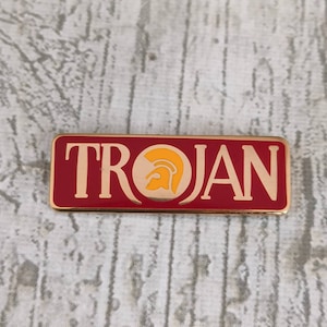 Puede incluir: Un pin de metal rojo y dorado con la palabra "TROJAN" en letras blancas y un gráfico de casco dorado.