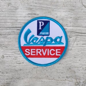 May include: A round, white patch with a blue border. The patch has a blue and white Vespa logo with the text "PIAGGIO" and the word "SERVICE" in red.