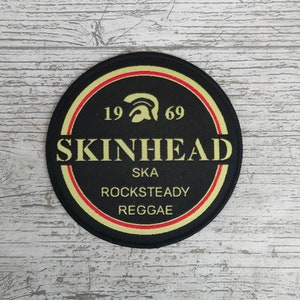 May include: A round black patch with the word "SKINHEAD" in yellow block letters. The patch also features the numbers "19 69" and the words "SKA ROCKSTEADY REGGAE". The patch has a yellow and red border.