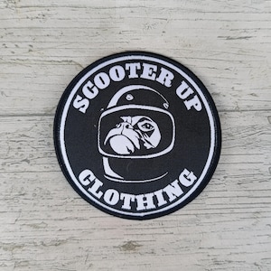 Peut inclure: Patch rond noir et blanc avec le texte "Scooter Up Clothing" en cercle autour d'un carlin de dessin animé portant un casque.