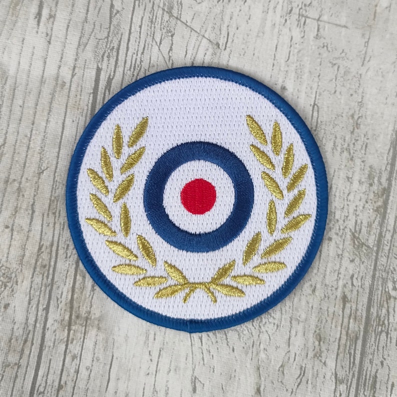 Puede incluir: Parche circular con fondo blanco, borde azul y dise&ntilde;o de diana en el centro. La diana tiene un punto rojo, rodeado de anillos azules. Hojas de laurel doradas bordadas alrededor de la diana. Un emblema decorativo.