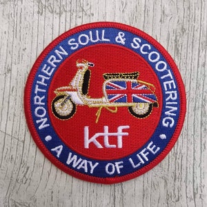 Puede incluir: Parche redondo bordado, con fondo rojo y borde azul. Presenta un scooter blanco con diseño de la Union Jack, y el texto "NORTHERN SOUL & SCOOTERING" y "A WAY OF LIFE". También se ven las letras "ktf".
