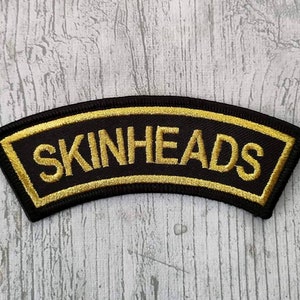 Puede incluir: Parche de tela negro con un borde bordado en oro y el texto "SKINHEADS" en oro.