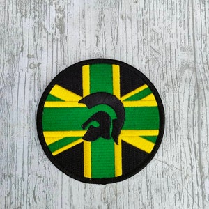 May include: A round black patch with a green, yellow, and black Union Jack design and a black silhouette of a Spartan helmet in the centre.