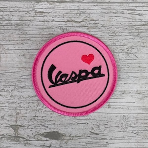 Puede incluir: Parche redondo bordado de color rosa con un logotipo de Vespa negro y un corazón rojo. El parche tiene un borde rosa.