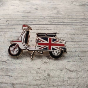 Puede incluir: Un pin de scooter blanco y dorado con un diseño de Union Jack en el lateral.