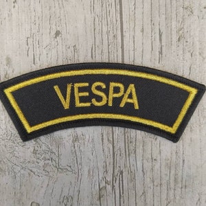 Puede incluir: Parche de tela negro con borde amarillo y la palabra "VESPA" bordada en amarillo.