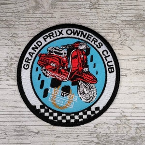 May include: A round, white and black embroidered patch with a blue background. The patch features a red scooter with a white and black chequered border. The text "Grand Prix Owners Club" is written in a white circle around the scooter.