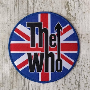 Könnte beinhalten: Ein runder, blauer, weißer und roter Patch mit dem Text "The Who" in schwarzen Buchstaben und einem nach oben zeigenden Pfeil. Der Patch zeigt ein Union-Jack-Design.