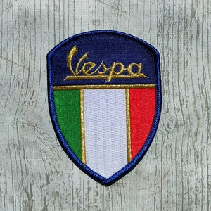 Puede incluir: Un parche bordado azul y dorado con la palabra "Vespa" y los colores de la bandera italiana.