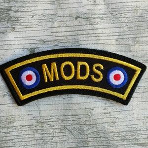 May include: Black embroidered patch with the word "MODS" in gold lettering. The patch has a gold border and features two roundels with red, white, and blue concentric circles, a classic design.