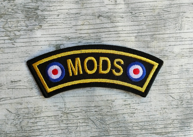 May include: A black embroidered patch with the word "MODS" in gold. The patch has a gold border and two roundels with red, white, and blue concentric circles. A classic design.