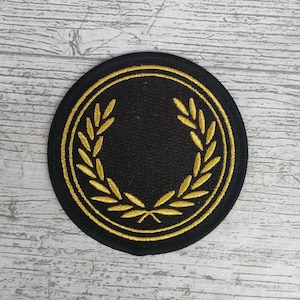 Może przedstawiać: Okrągła, czarna naszywka ze złotym wzorem wieńca laurowego. Naszywka ma złote obramowanie i prawdopodobnie jest haftowanym emblematem, odpowiednim do przyszycia do odzieży lub akcesoriów.