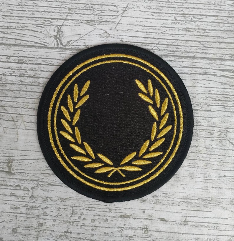 May include: A round, black patch with a gold laurel wreath design. The patch has a gold border and is likely an embroidered emblem, suitable for sewing onto clothing or accessories.