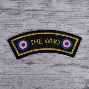 Puede incluir: Parche bordado negro y dorado con el texto "THE WHO" y dos símbolos de diana rojo, blanco y azul.