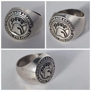 May include: A silver ring with a circular design featuring the text "Skinheads Against Racial Prejudice" and a stylized image of a Spartan helmet with the word "SHARP" above it.