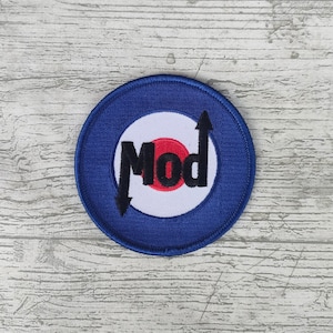 Puede incluir: Un parche redondo azul, blanco y rojo con la palabra "Mod" en negro. El parche tiene un diseño de diana con flechas que apuntan hacia arriba y hacia abajo. Ideal para ropa o accesorios.