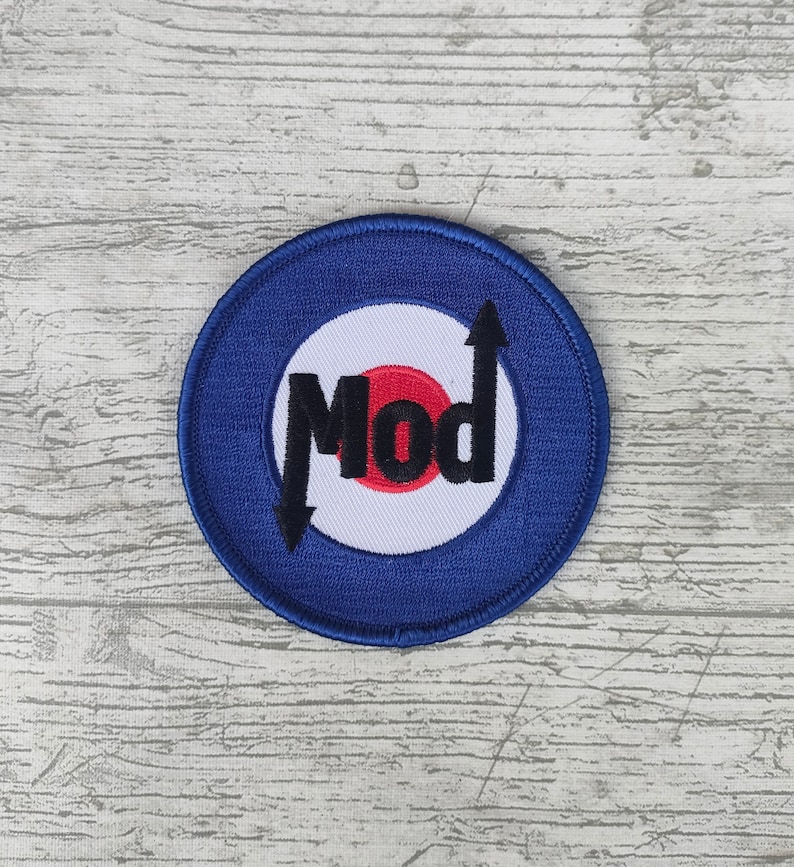 May include: A round, blue, white, and red patch with the word "Mod" in black. The patch has a target design with arrows pointing up and down. The patch is likely for clothing or accessories.