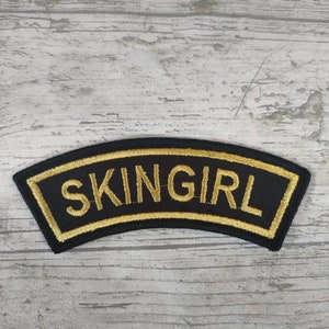 May include: Black and gold embroidered patch with the word "SKINGIRL" in a curved shape.