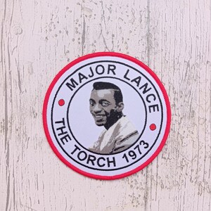 May include: A white and red circular patch with a black and white photo of Major Lance. The text on the patch reads "Major Lance The Torch 1973".