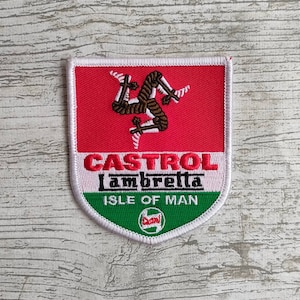 Puede incluir: Un parche bordado blanco, rojo y verde con el texto "Castrol Lambretta Isle of Man" y una imagen estilizada de la bandera de la Isla de Man.