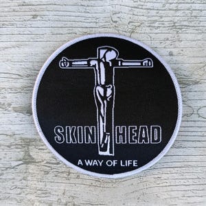 Puede incluir: Un parche bordado circular de color negro con un borde blanco. El diseño presenta el contorno blanco de una figura en una cruz, con el texto SKIN HEAD a ambos lados de la cruz y el texto A WAY OF LIFE impreso en color blanco debajo.