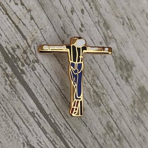 Peut inclure: Épinglette en métal doré représentant une figure stylisée d'une personne avec les bras tendus, portant une tenue noire et bleue, sur une croix. La figure a une tête blanche avec un contour noir.