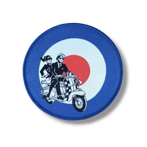 May include: A circular patch with a blue border and a white, red, and blue target design. The patch depicts a black and white illustration of a couple on a scooter. The scooter has a checkered pattern.