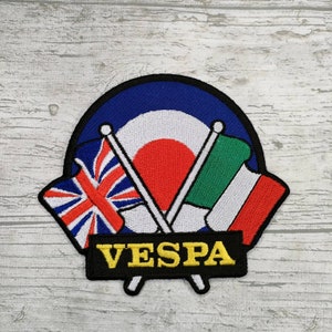 Puede incluir: Un parche bordado en blanco y negro con un blanco, rojo y azul en el centro. El parche presenta dos banderas cruzadas, una con la Union Jack y la otra con la bandera italiana. El texto "VESPA" está escrito en letras amarillas sobre una pancarta negra.
