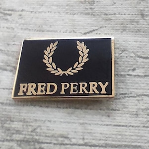Może przedstawiać: Złota i czarna emaliowana szpilka z wieńcem laurowym i napisem "FRED PERRY".