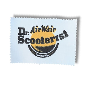 May include: A rectangular white cleaning cloth with pinked edges featuring a black and yellow graphic logo. The text reads Dr. AirWair Scooterist with a circular emblem containing the words With Scooter Up Soles in a black and yellow design.