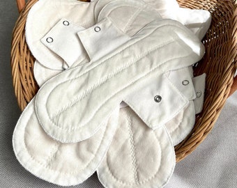 May include: A selection of off-white, reusable menstrual pads with popper fastenings, presented in a woven wicker basket. The pads feature a quilted design and are made of soft, absorbent fabric. The basket has a natural wood finish and a curved handle.