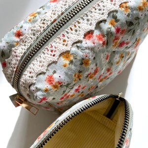 May include: A quilted floral makeup bag with a white lace trim and a zipper closure. The bag is open to reveal a yellow and white striped lining.