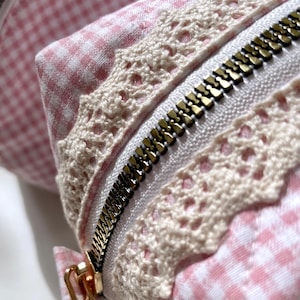 May include: A pink and white gingham fabric makeup bag with a gold zipper and white lace trim.