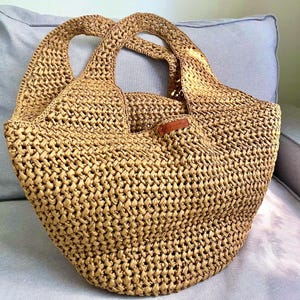 May include: A woven straw tote bag with two rounded handles. The bag is a light tan color and has a woven pattern. A small brown leather tag is attached to the bag. The bag is sitting on a light blue surface.
