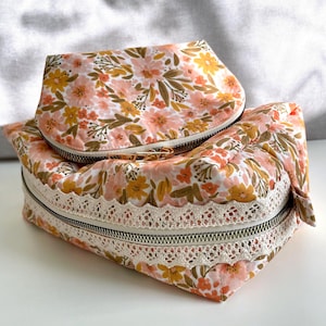 May include: Two floral print zippered pouches with a white lace trim. The smaller pouch is round and sits on top of the larger rectangular pouch.